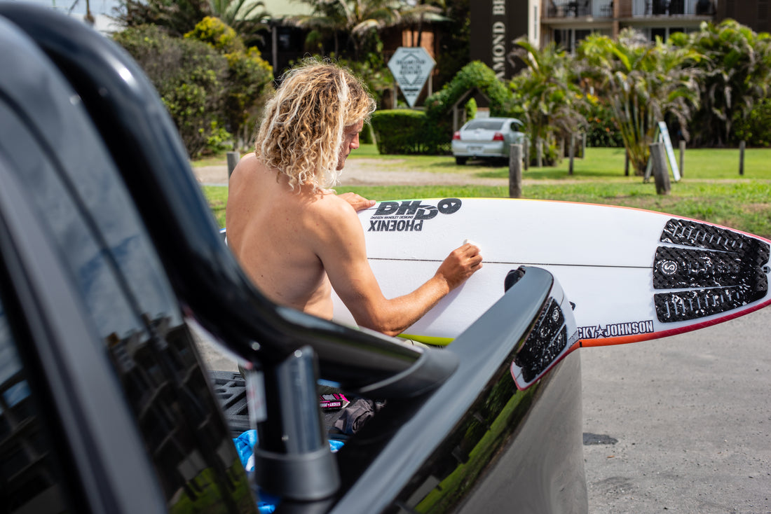 A surfer waxing a shortboard on the back of a ute before a surf