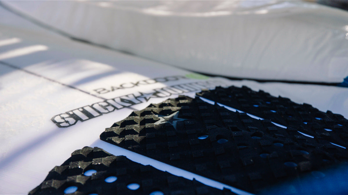 A black Sticky Johnson three-piece tail pad on the tail of a white surfboard