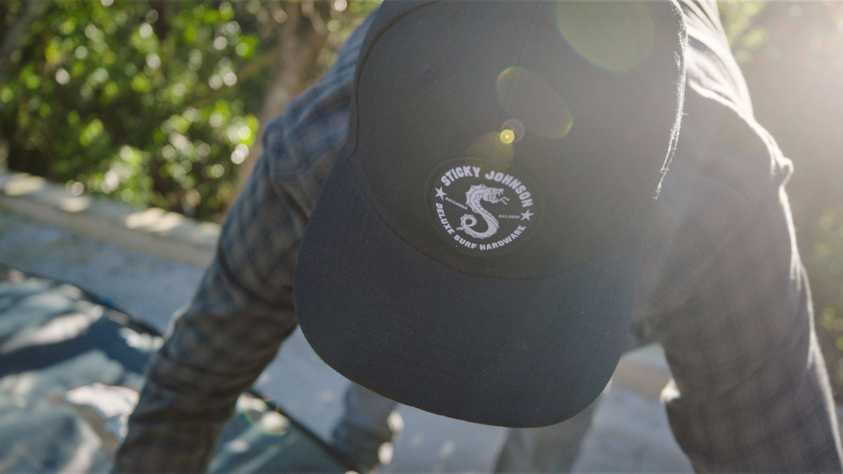 A surfer in a Sticky Johnson cap leaning over a board in the morning sun